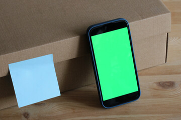 Smartphone with green screen leaning on cardboard box with sticky note