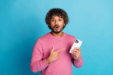 Young man with a surprised expression pointing to a smartphone in his hand, standing against a...