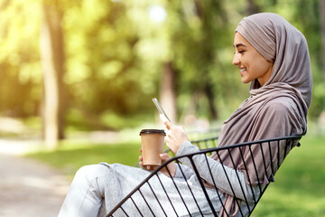 Profile photo of arab woman drinking coffee and using phone, sitting on bench at park, empty space