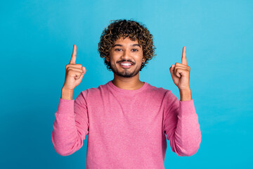 Cheerful Young Man Gesturing Upwards Against a Vibrant Blue Background, Wearing a Casual Pink Sweater and Smiling Brightly