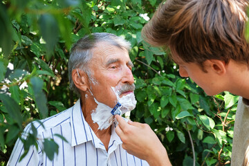 A young man carefully shaves an elderly man's face outdoors, surrounded by green foliage.