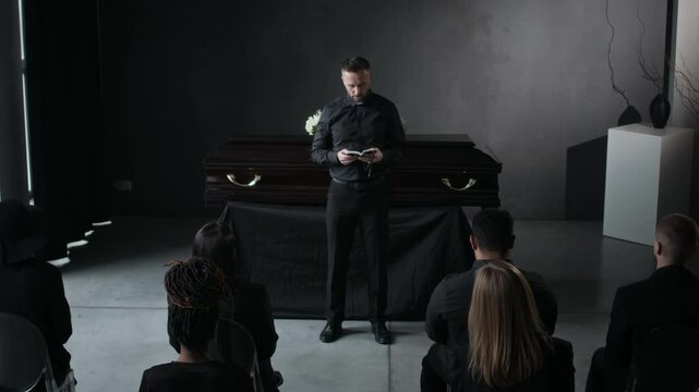 High angle shot of man in black delivering eulogy near white-flowered coffin before several sitting mourners in dark clothing at funeral