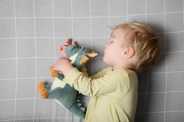 Sleeping Toddler Hugging Plush Dinosaur on Grey Grid Bed