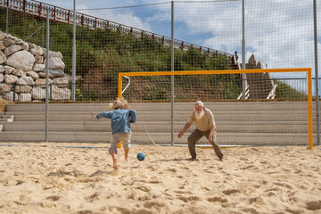 Barefoot child kicking soccer ball towards elderly man acting as goalkeeper on sandy beach field, concept of intergenerational activity, family bonding, summer sport and outdoor fun.