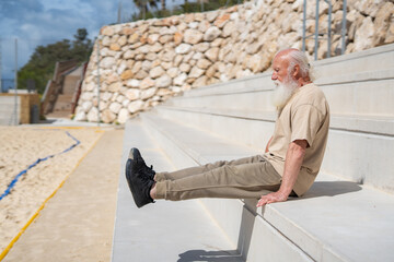Active senior man performing seated leg lift exercise on concrete steps outdoors, demonstrating...