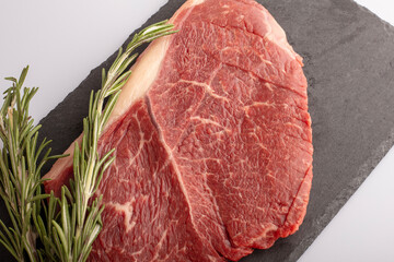 A thick ribeye steak with rich marbling is placed on a dark slate board alongside fresh rosemary sprigs, ideal for grilling or roasting. Perfect for a delicious meal