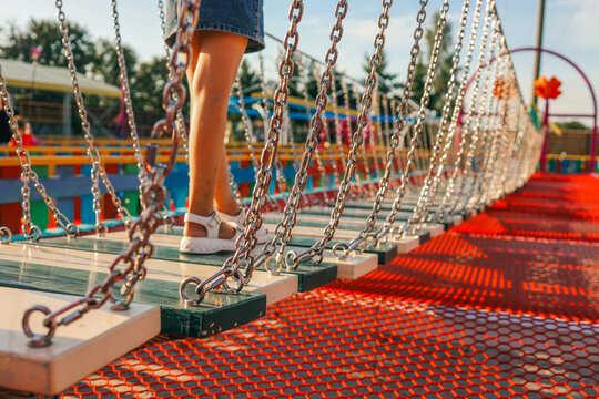 Child crossing rope bridge on playground. Legs in white sandals walking on suspended wooden planks, colorful background with chains and net shadows