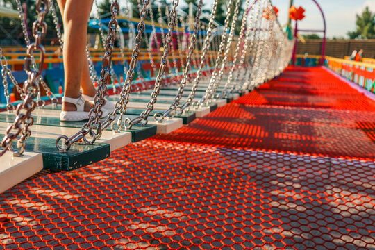 Fototapeta Child crossing rope bridge on playground. Legs in white sandals walking on suspended wooden planks, colorful background with chains and net shadows