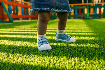 Toddler in blue sneakers standing on bright green artificial grass in a colorful playground. Low angle view of children's legs on a sunny day