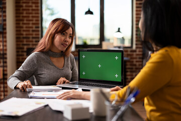Laptop displaying greenscreen as asian businesswoman speaks to her coworker at desk, discussing marketing strategies. Female colleagues share project plans, using digital device with mockup template.