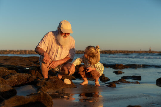 Grandfather showing seashell to curious grandson on beach at sunset, bonding moment during seaside exploration.