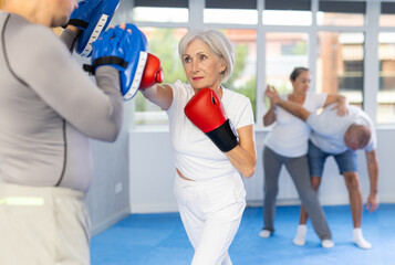 Sportive mature female practitioner of boxing courses applying kicks on hitting mitts during...