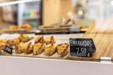 Close-up of golden baked empanadas inside a takeaway counter, with a price tag reading 2.50 euros. Blurred background, focus on product. Horizontal image, eye-level shot.
