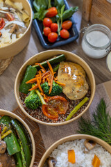 Overhead view of assorted takeout bowls with grilled chicken, vegetables, quinoa, pasta, and rice. Fresh herbs, tomatoes, and glass jar add detail. Colorful food styling. Vertical, copy-space.