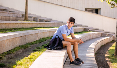 Teenage boy student 17 years old with a backpack sits on the wooden steps outside the college