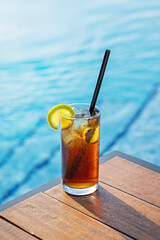 Cuba Libre cocktail with ice, a lemon slice, and a straw on a wooden table near a blue pool.