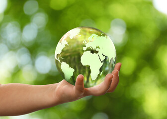 Child holding glass ball with Earth globe inside on blurred green background, closeup. Concept of environment protection