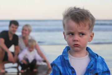 portrait of serious toddler by the ocean with his happy family in background