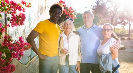 Portrait of happy multiracial various aged adult people bonding to each other and looking at camera while standing against the background of flowering and green trees in city park on a sunny day