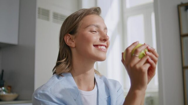 Woman biting tasty apple in sunny kitchen. Closeup girl admiring fresh fruit