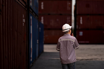 A logistics supervisor in a hard hat and uniform communicates via radio while holding a clipboard...