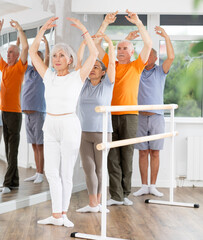 Fototapeta premium Beautiful mature women and sports men practice at ballet standing in fifth position at barre in bright choreography studio
