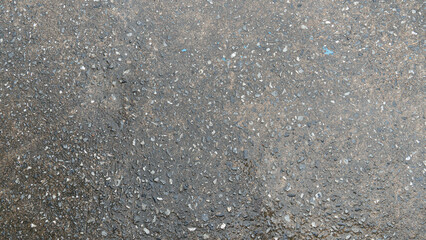 Textured surface of asphalt road. Rough grainy texture is visible with small pebbles or aggregate embedded in the gray material. Focus is on the surface texture and detail of water from rain on top.