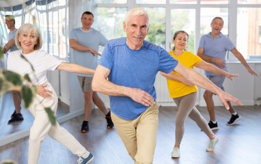 Expressive cheerful gentleman participating in upbeat Zumba class for group of seniors in mirrored...