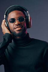 Obraz premium Young Black man in stylish black turtleneck and sunglasses enjoying music with headphones, expressing happiness and confidence against a gradient background