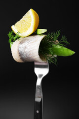 Fork with marinated herring fillet, cucumber, dill and lemon on black background, closeup