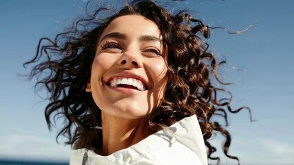 A joyful woman with curly hair smiles at the sky by the sea. Captured in a low-angle shot, the video conveys freedom and happiness.