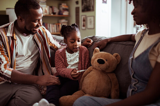 Young african american parents teaching their daughter how to use a thermometer at home on the couch