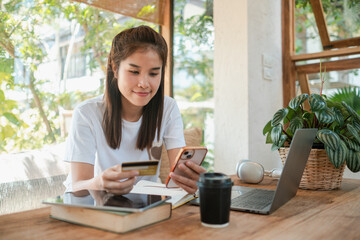 A young woman uses her smartphone and credit card for online shopping at a home office desk with a laptop and coffee.