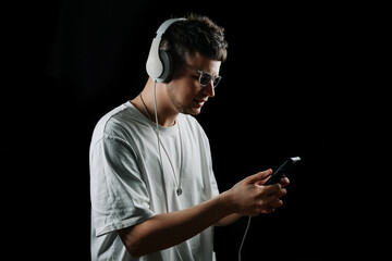 Naklejka premium Young man in white t-shirt and headphones looking at smartphone, listening to music or podcast. Isolated on black background. Modern digital lifestyle.