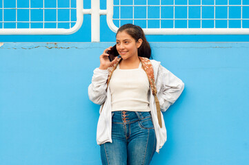 A young woman is happily conversing on her mobile phone while leaning against a vibrant blue wall,...