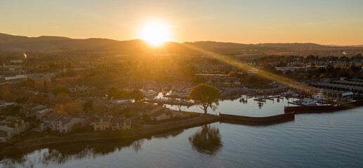Aerial view of a marina in Pittsburg, California, USA, at sunset. Boats are docked, reflecting in the water, with houses and hills in the background. The sun is setting over the horizon.