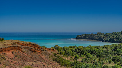 Beautiful serene seascape. A calm turquoise ocean, waves foaming at the shore. Lush green vegetation and a red-soil hill in the foreground. Clear blue sky. Copy  space. Madagascar. Nosy Iranja   