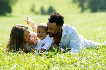 Multi racial parents and mixed race baby in park. Multiethnic family outdoor. Biracial baby child on yard. Diverse Multi racial family. Multi racial father and mother with child lying on grass.