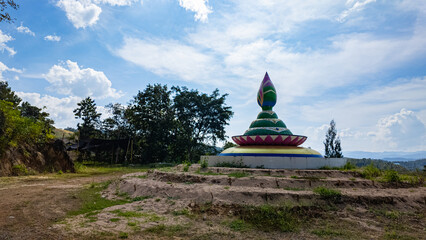  A vibrant stupa in the shape of a lotus, featuring intricate green, pink, and yellow details, set against a bright blue sky with scattered clouds. Surrounded by a serene natural landscape