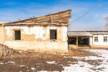 This image captures a mud-brick building with a partially collapsed wooden roof and broken windows. The house sits in a rural setting with patchy snow and dry ground highlighting the harsh environment