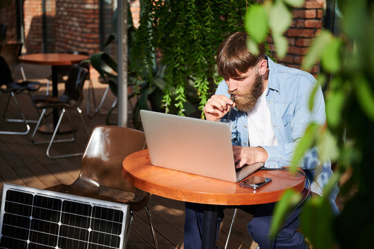 Man works on laptop and smartphone at outdoor table, with glass of iced coffee. Solar panel nearby, emphasizing sustainable, modern eco-friendly workspace that combines technology and sustainability. - Powered by Adobe