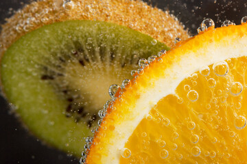 Pieces of tropical fruits of fresh orange and green kiwi in water with air bubbles on a black background. Macro.