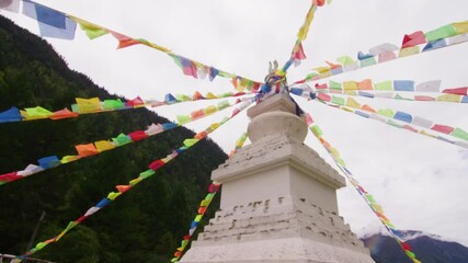 A white stupa in Jiuzhaigou is crowned with radiating Tibetan prayer flags, set against a cloudy sky and forested mountains.