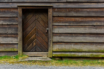 A rustic wooden door featuring a chevron design stands sturdily against an old log cabin on a cloudy day with grassy ground.