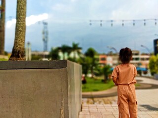 Medan/North Sumatra-April 1 2025: a little girl is enjoying the view amidst the hustle and bustle of the city park.child,back,park,city,road,adventure,alone,calm,relax,clothes,sand,tree,leaves,sky,clo