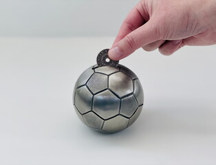A person’s hand deposits a coin into a soccer ball piggy bank, symbolizing saving money. The coin...