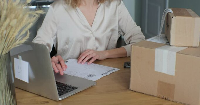A young woman writes a return order and sits at a table with a laptop at home. Close-up video of a female customer writing on paper and filling out a form, wanting to return a product.