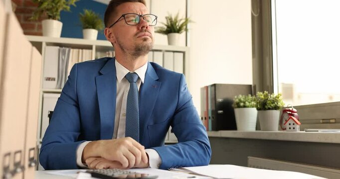 Thoughtful man worker in glasses sits at desk in office. Serious working clerk in jacket reflects on tasks during working shift near window in room