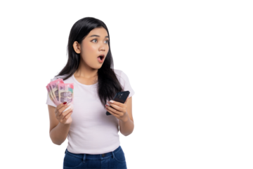 Surprised young Asian woman holding Indonesian rupiah cash in one hand and smartphone in the other, looking away, reacting to unexpected financial news, isolated on transparent background