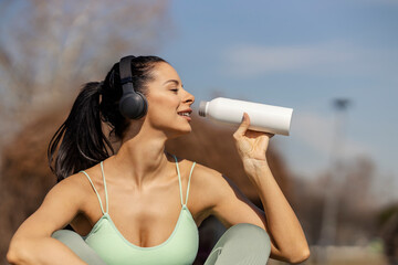 Portrait of thirsty sportswoman sitting on outdoors and refreshing herself with bottle of water.